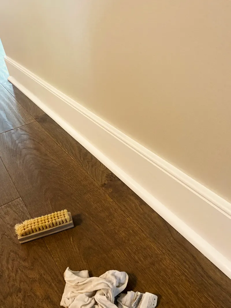 Painted wall and white baseboard meeting a hardwood floor, with a brush and rag